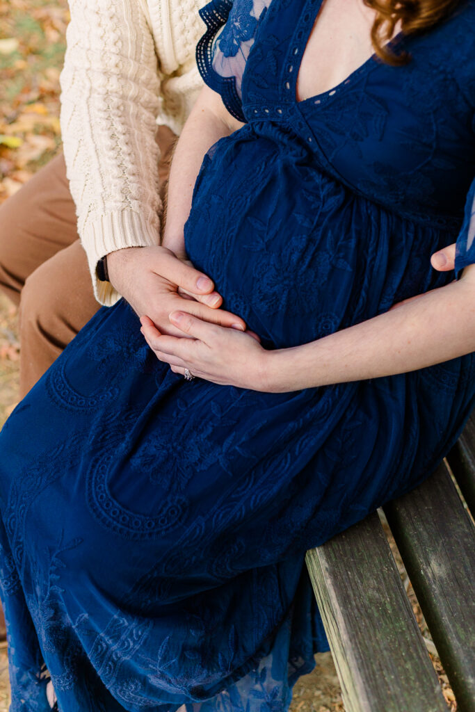Beautiful mama to be is sitting next to her husband as both are holding her baby bump in a outdoor Pittsburgh, PA Maternity Photography session in the fall.