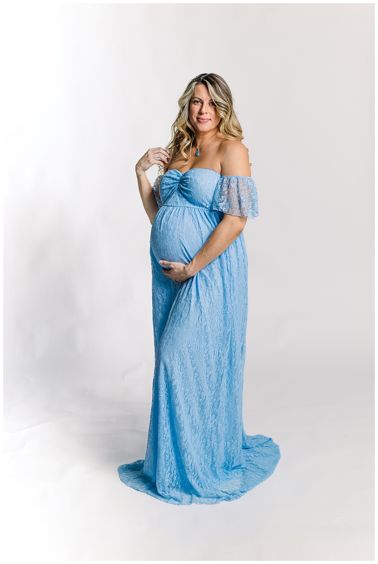 Mom holding baby bump in a beautiful off the shoulder blue dress during her indoor studio session while a Maternity Photographer in Pittsburgh, PA took her maternity pictures