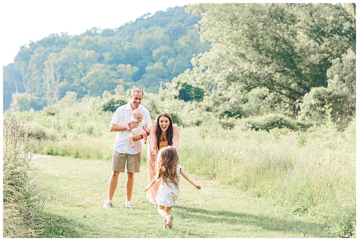 Mom and Dad are holding baby brother while the young girl is running towards them. This is taken in a wildflower field, during a pittsburgh family session.