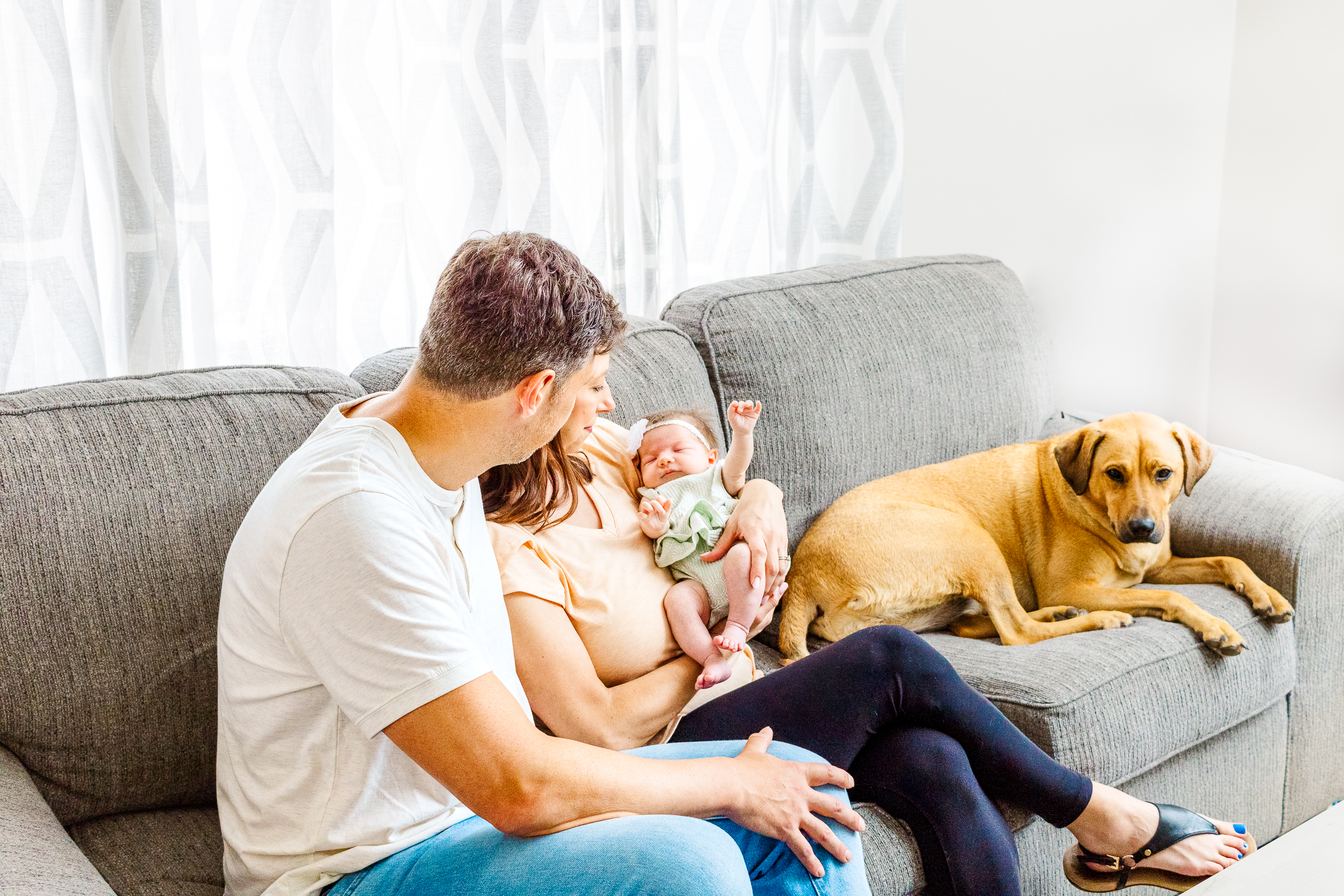 Parents cuddling their newborn baby on the couch in their living room, captured in a calm, relaxed lifestyle session in Pittsburgh.