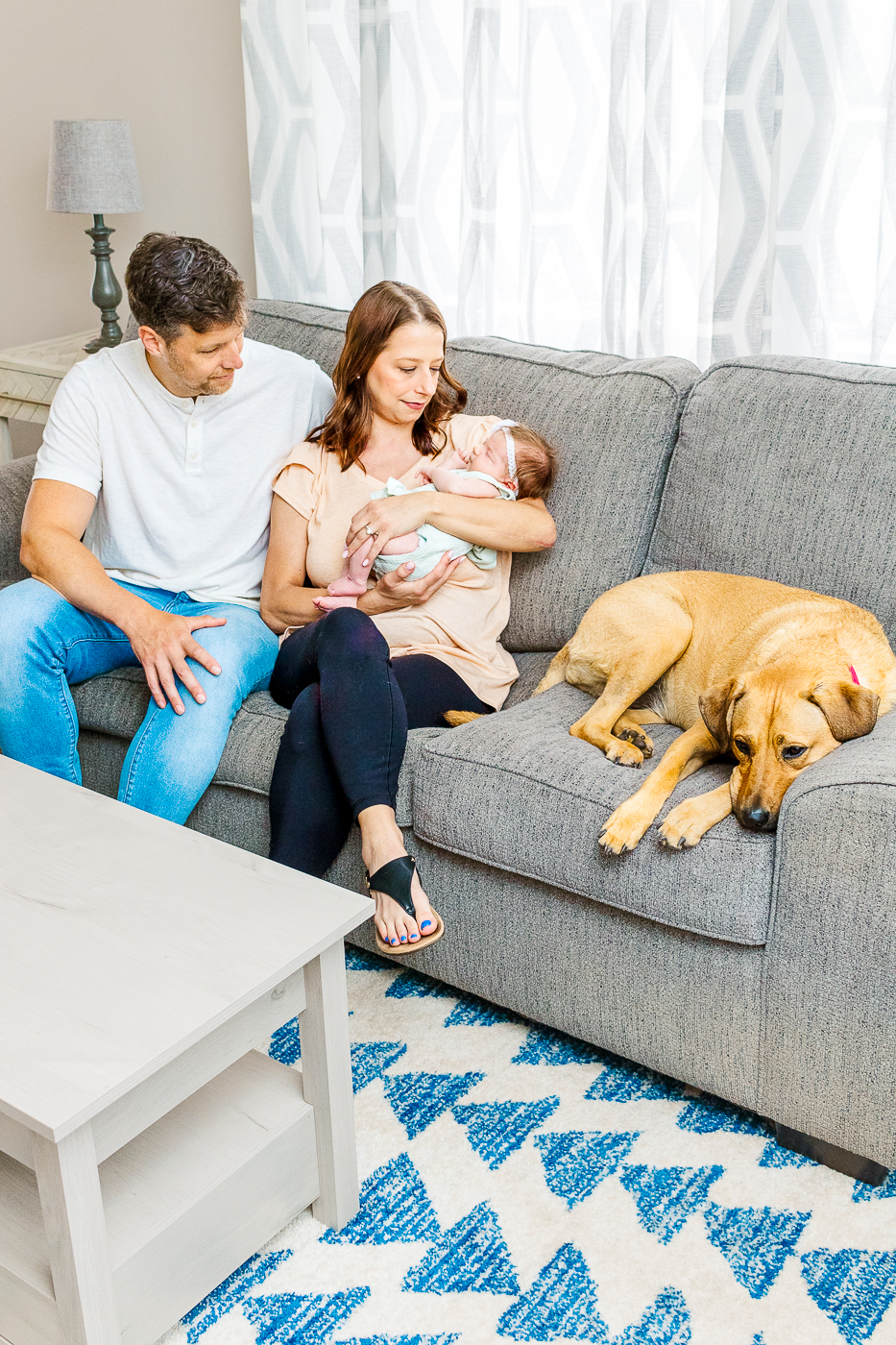 Mom is holding baby girl while dad is sitting next to them and their fur baby is laying on the couch next to them. This is part of the 9 real life tips to help you prepare for your baby in wexford.