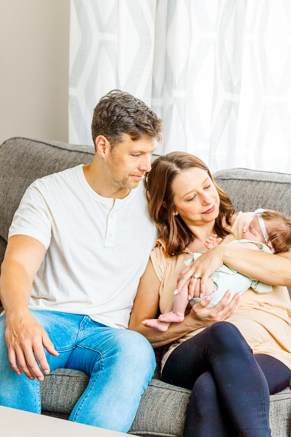 Parents cuddling their newborn baby on the couch in their living room, captured in a calm, relaxed lifestyle session in Pittsburgh.