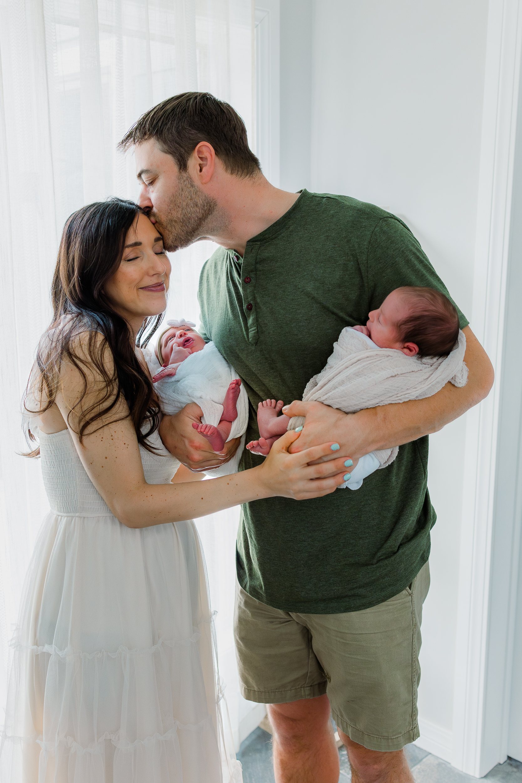 Mom and Dad are holding their newborn twins. Dad is kissing mom on the head. Sweet moment taken during a lifestyle in home newborn session in Pittsburgh.